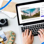 Overhead view of hands typing on a laptop to book a Paris trip online, surrounded by a camera, map, and travel accessories on a white desk.
